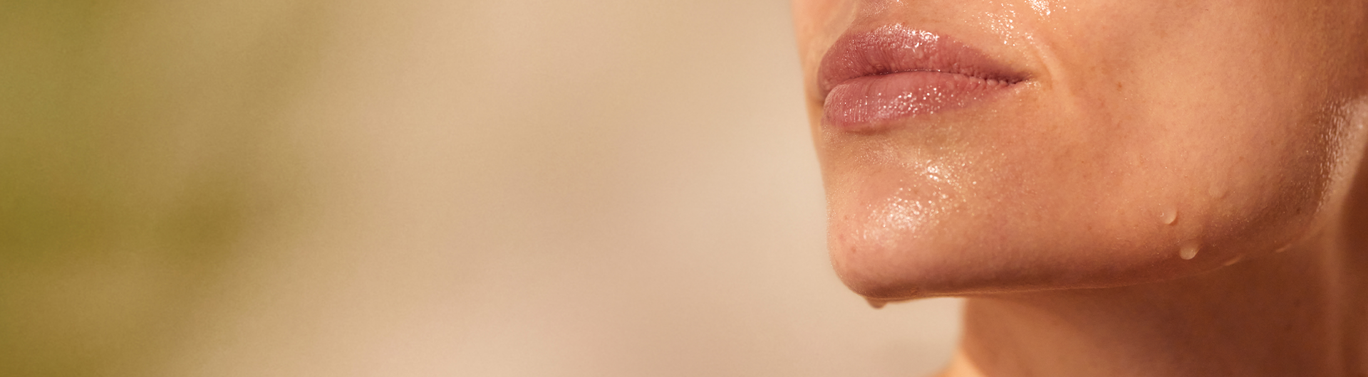 Shadow and beige background and to the right lower part of a woman's face. The skin is hydrated with droplets of water on the lower jawline and on the chin. The skin surface is glowy in the light.